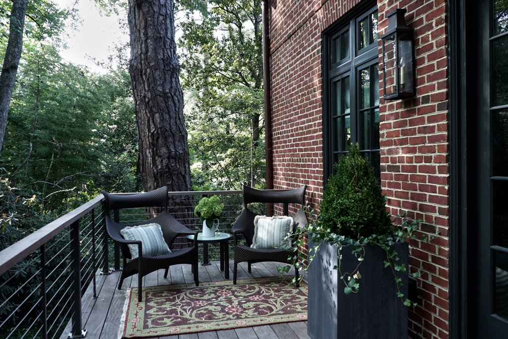 A cozy outdoor balcony patio with two black chairs, a small round table, a potted plant, and cushions, with a brick wall and large window behind.