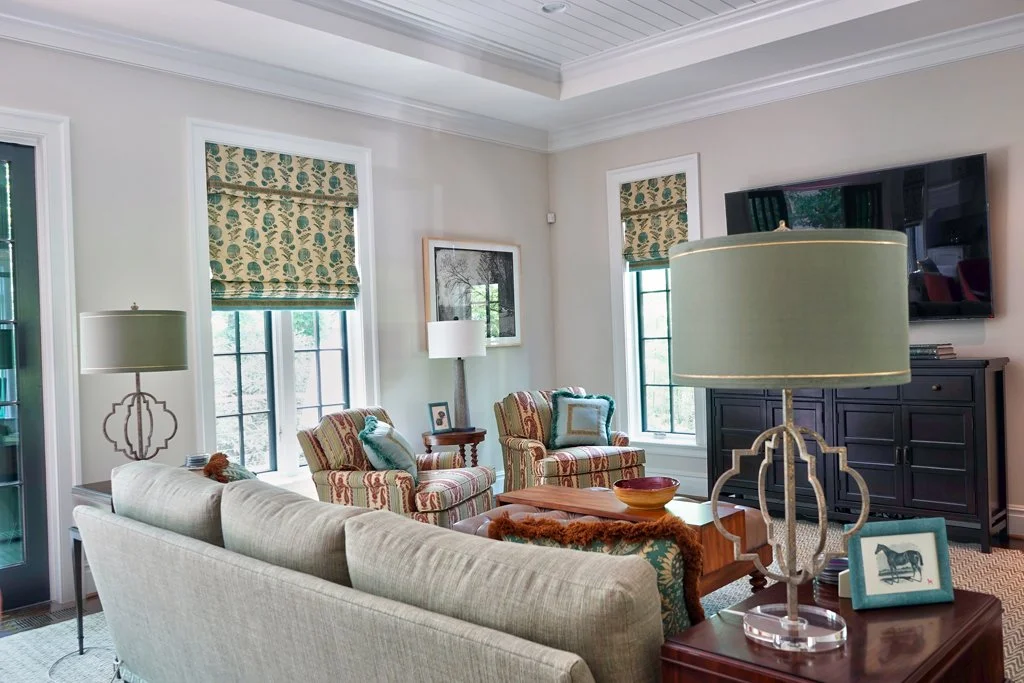 Living room with beige sofa, two striped armchairs, side table, wall-mounted TV, framed artwork, window blinds with a leaf pattern, and various lamps.