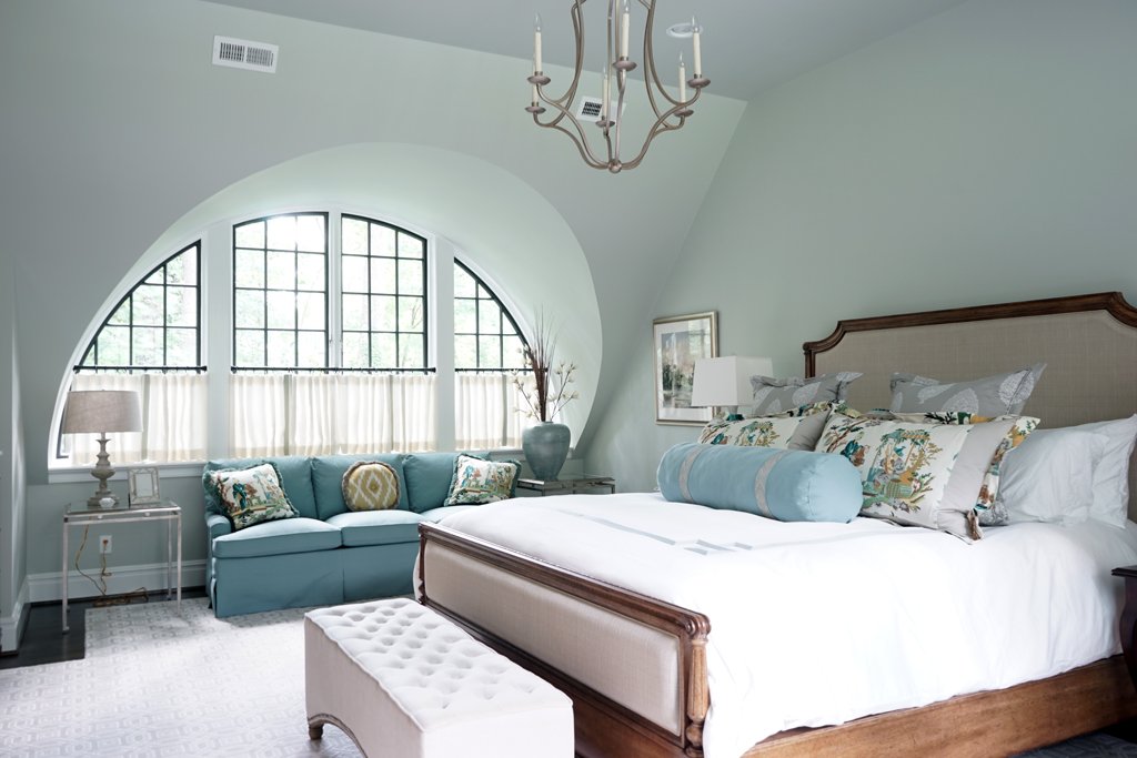 A bedroom with a large arched window, a beige upholstered bed with decorative pillows, a blue bench at the foot of the bed, a side table with a lamp, a chandelier, and a window seat with cushions and pillows.