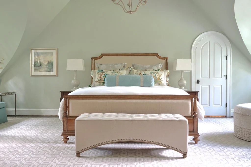 A bedroom with a wooden bed, light-colored upholstered headboard, and a long decorative pillow. There are two matching nightstands with lamps, a framed picture on the light green walls, an arched white door, and a cream-colored upholstered bench at t