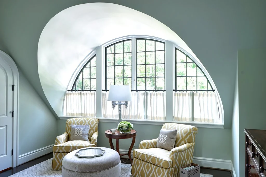 A cozy sitting area with two yellow patterned armchairs, a round ottoman, a wooden side table with a lamp and flowers, and a large arched window with white curtains in a room with light walls.