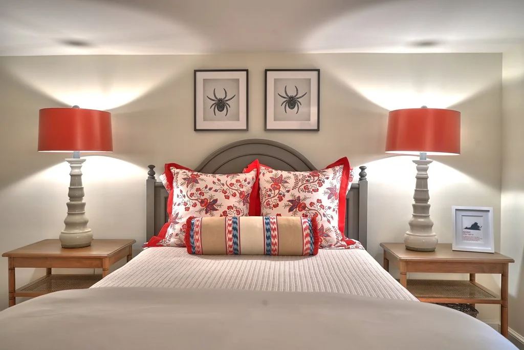 A neatly made bed with white bedding, red and white floral pillows, a long beige and red striped pillow, flanked by two wooden nightstands with table lamps with red shades. Two framed pictures of spiders are hanging above the headboard, which is gray