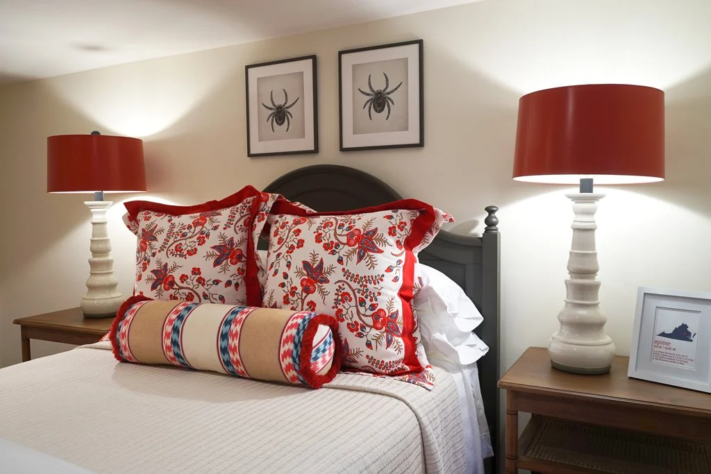 A bedroom with a bed that has a dark gray headboard, decorated with embroidered pillows, and a long cylindrical bolster pillow. Two matching white lamps with red shades are on wooden nightstands, along with framed pictures on the wall and a framed an