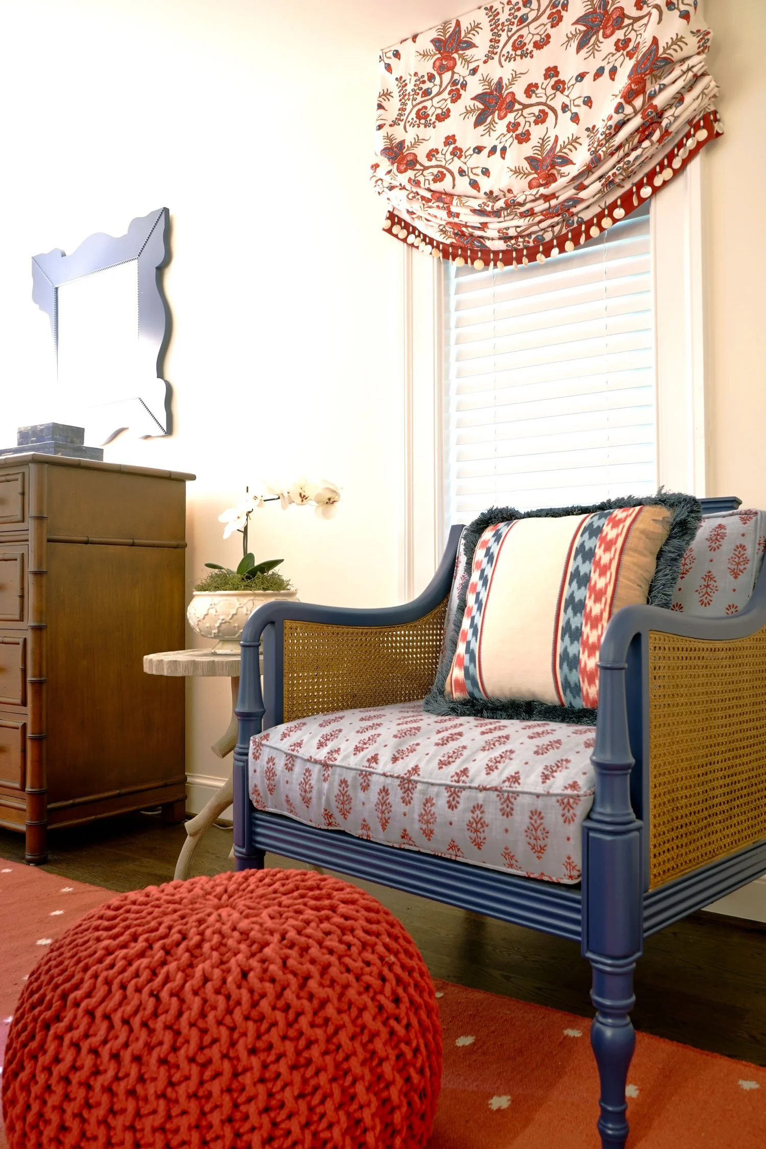 A cozy living room corner with a blue-framed vintage-style loveseat with patterned cushions, a small side table with a white orchid, a wooden chest of drawers with a mirror above, a window with a floral valance, and a large red knitted pouf on a burn