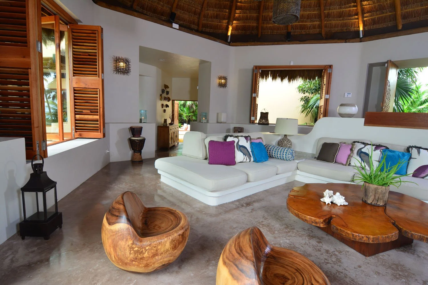 Interior view of a tropical living room with white L-shaped sofa, colorful pillows, wooden furniture, open windows with wooden shutters, and lush greenery outside.