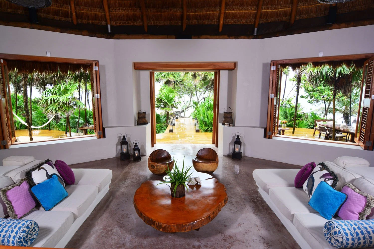 Living room with open windows revealing lush green tropical outdoor scenery, featuring wooden furniture and colorful cushions.