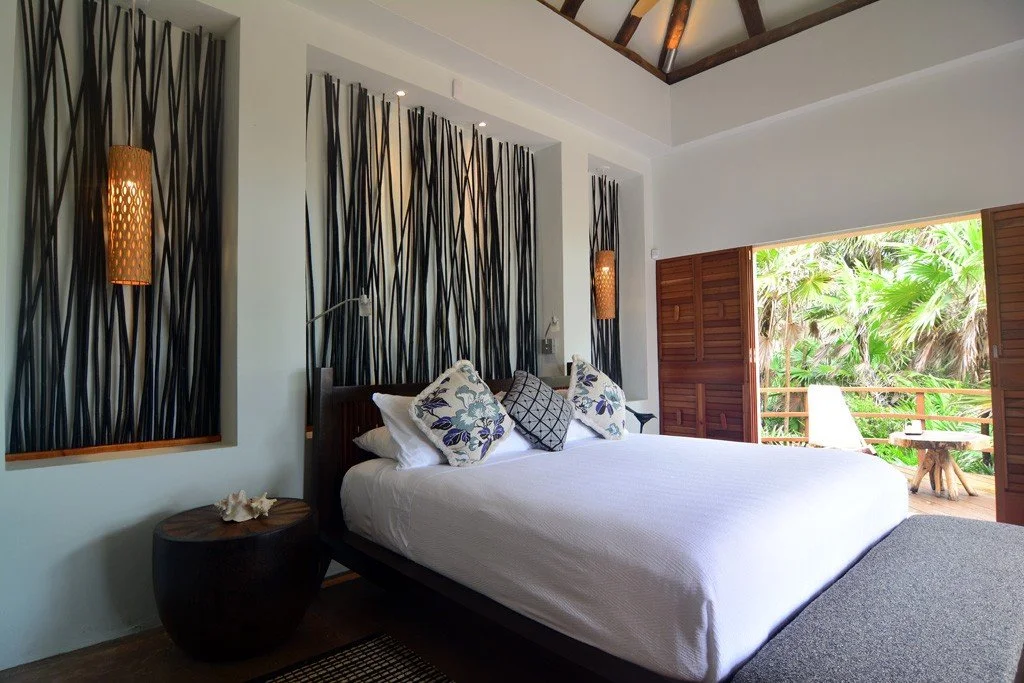 Hotel room with a large bed, decorative pillows, wall-mounted lamps, a small side table with seashells, and an open balcony with tropical greenery outside.