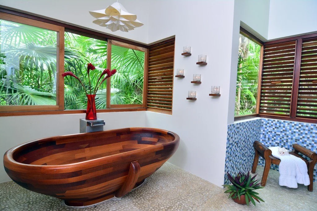 A bathroom with a wooden bathtub, large windows showing green tropical plants, a vase with red flowers, wall-mounted candles, a small seating area with towels, and wooden louvered window panels.