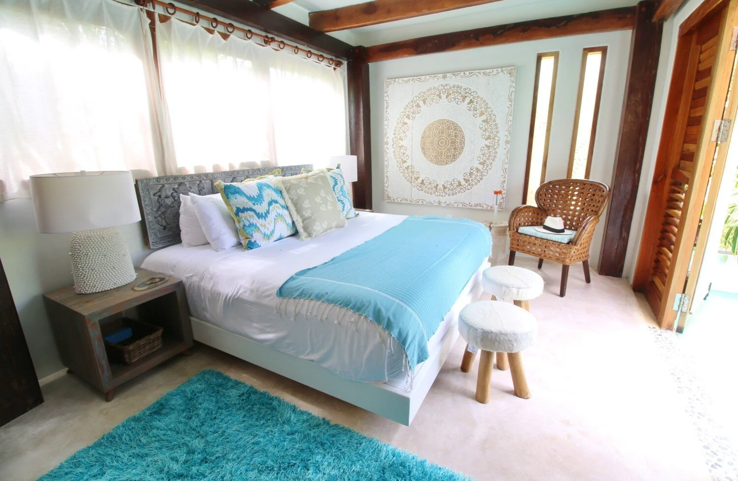 Cozy bedroom with a large bed, blue and white bedding, decorative pillows, two white fluffy stools, rattan chair with hat, nightstand with lamp, large window with sheer curtains, textured wall art, wooden beams, and open door with greenery outside.
