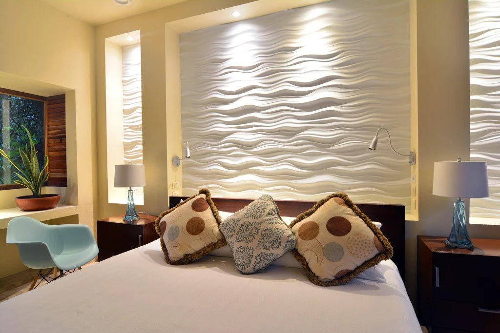 Modern bedroom with a white textured accent wall behind the bed, two nightstands with lamps, a window with blinds, and decorative pillows on the bed.