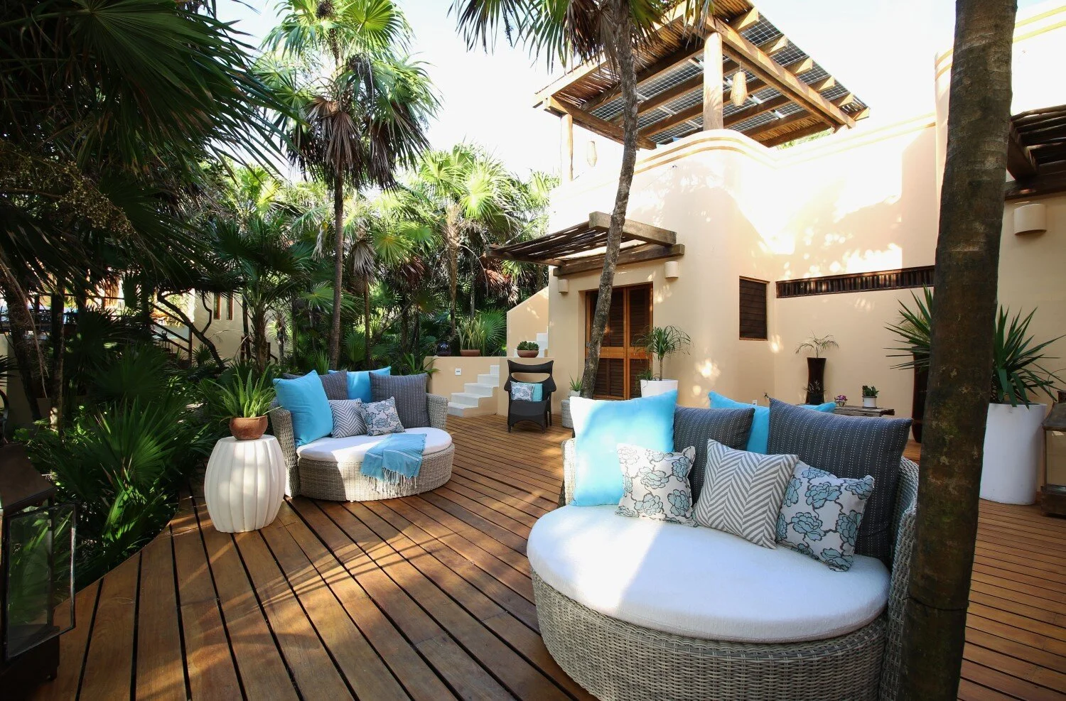 Outdoor patio with wicker sofas and chairs with blue and patterned cushions, potted plants, lush green trees, and a wooden deck, near a beige modern house with wooden accents and shaded areas.