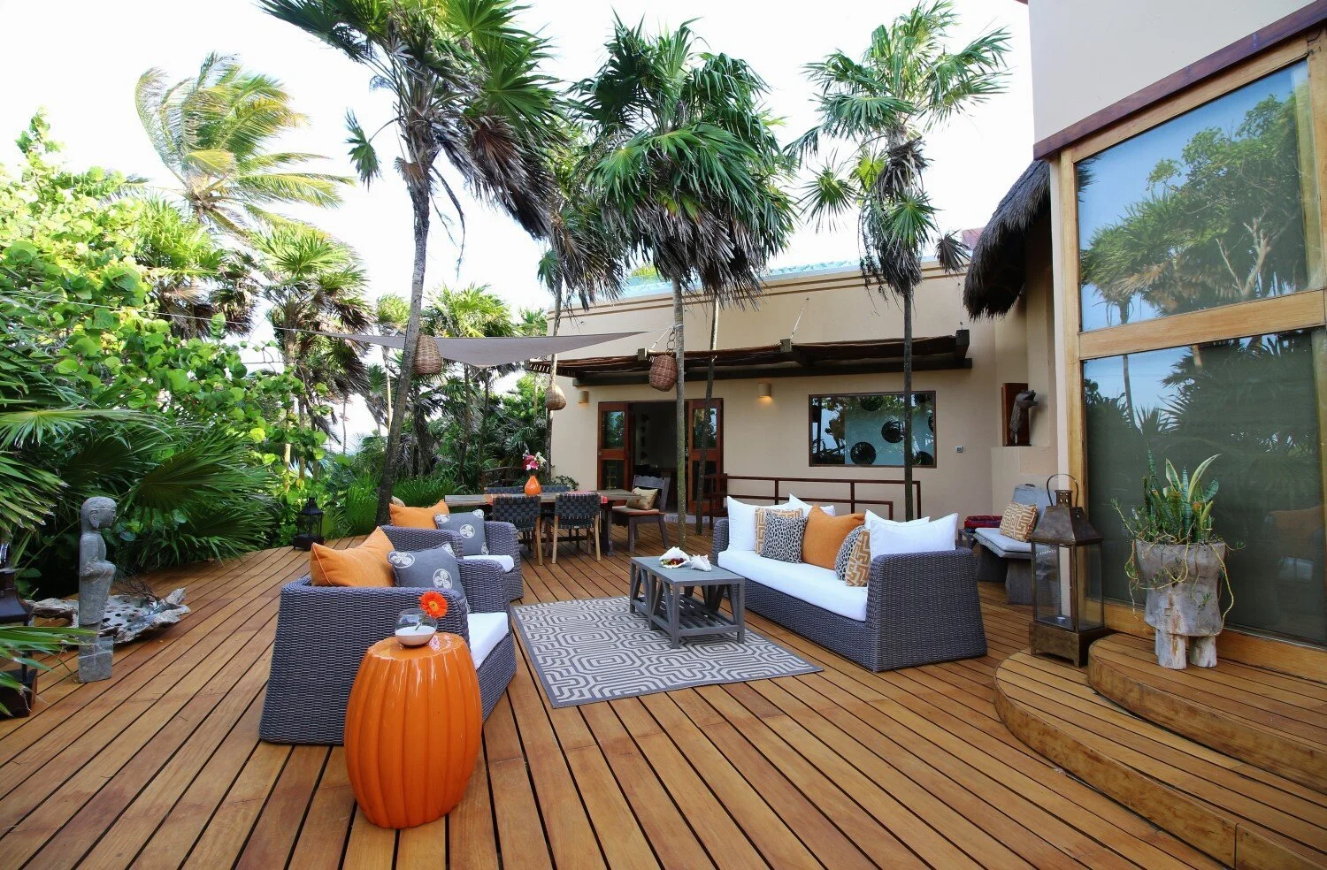 Outdoor patio with wicker furniture including a couch and chairs, decorated with colorful pillows, on a wooden deck surrounded by tropical trees and plants, with a house in the background and blue sky.