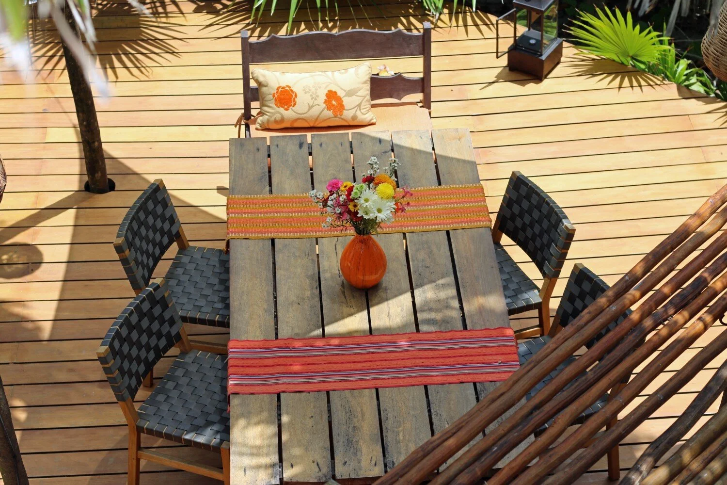 A wooden outdoor table with a central orange vase holding a mixed flower bouquet. There are two orange and red striped table runners across the table. Six woven black chairs are around the table. In the background, a lounge chair with an orange decor