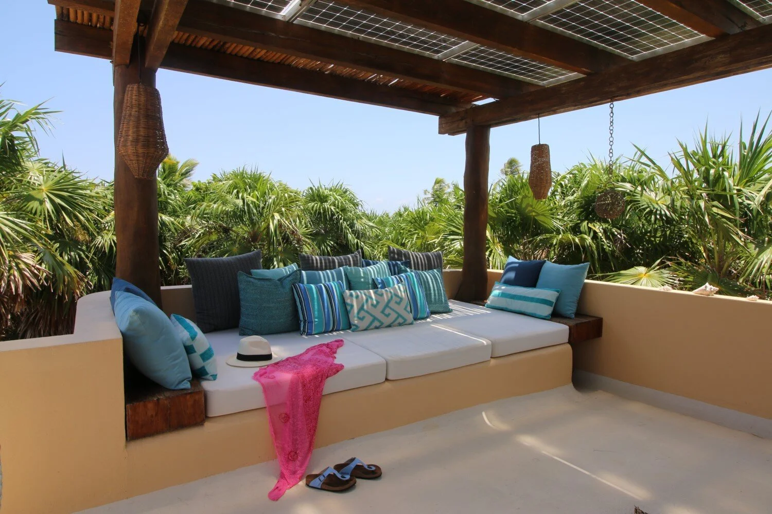 A cozy outdoor patio with a built-in bench, decorated with blue and teal pillows, a white hat, a pink towel, and a pair of sandals, surrounded by lush green tropical plants under a wooden roof with hanging wicker lanterns.