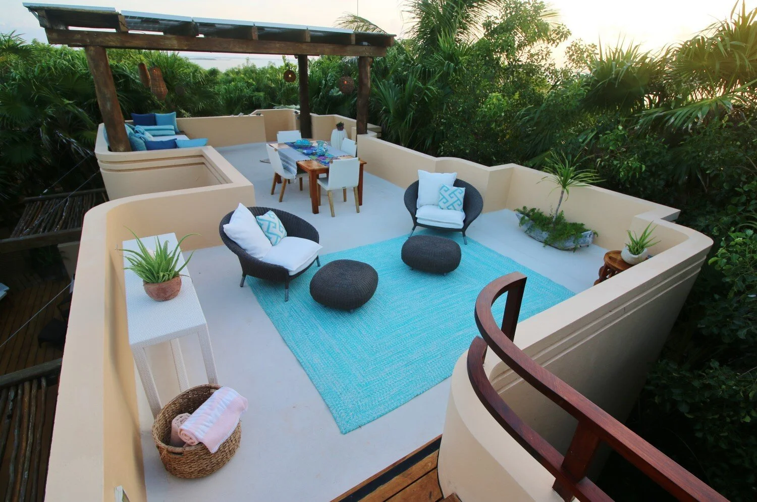 Outdoor terrace with beige walls, featuring two black wicker chairs with white cushions and blue patterned pillows, a blue area rug, and small potted plants. There is a dining table with four chairs, and a shaded corner with blue cushions and a woode