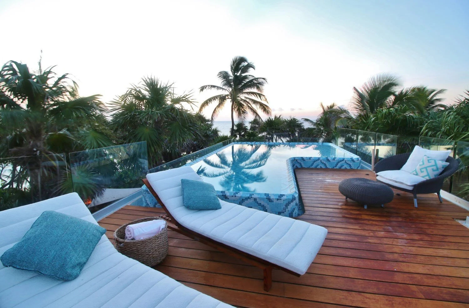 View of an outdoor rooftop pool area with lounge chairs and tropical palm trees, overlooking the ocean at sunset.