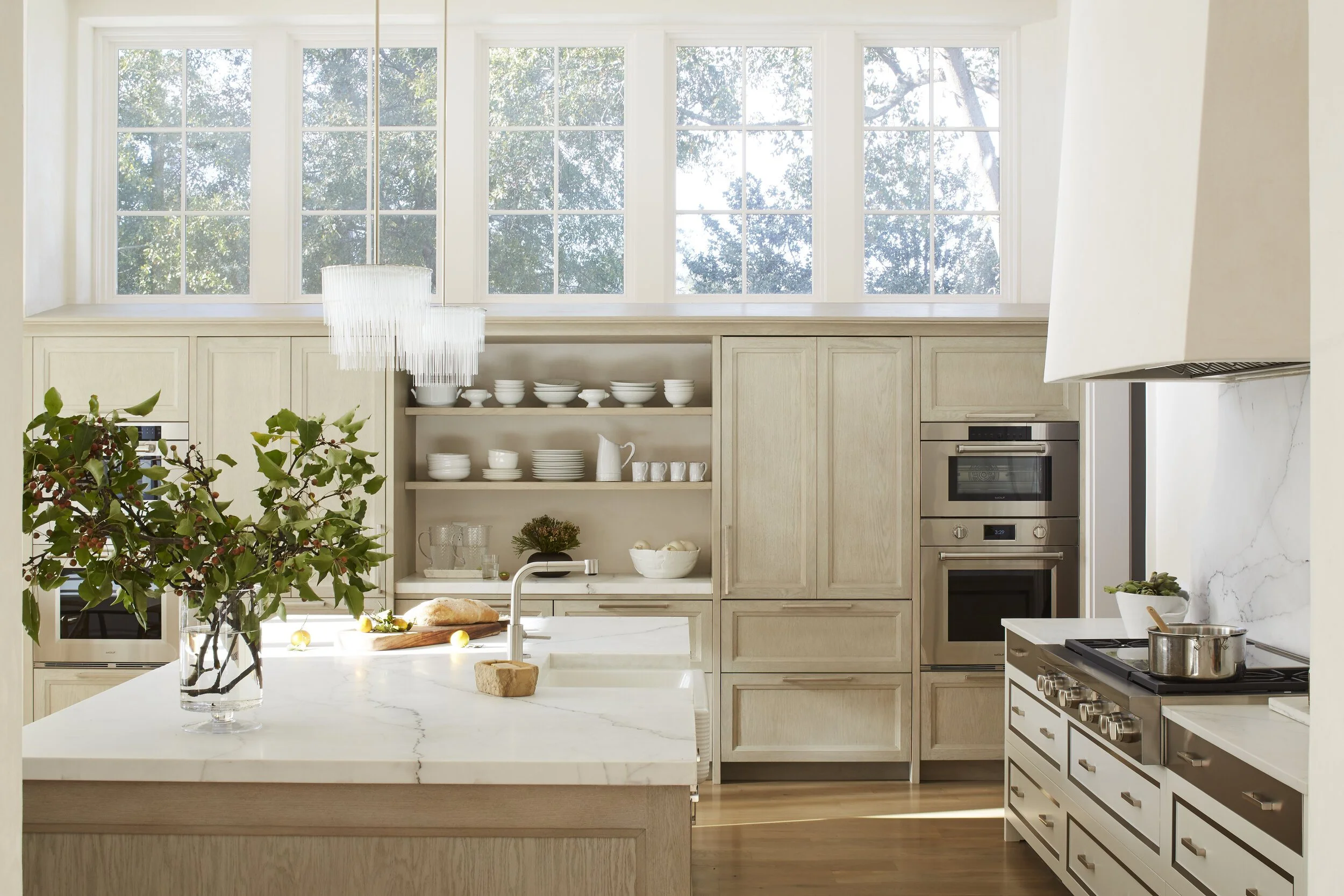 Bright kitchen with white cabinetry, a marble countertop island, open shelves with white dishes, a large window letting in natural light, green plants, and modern appliances.