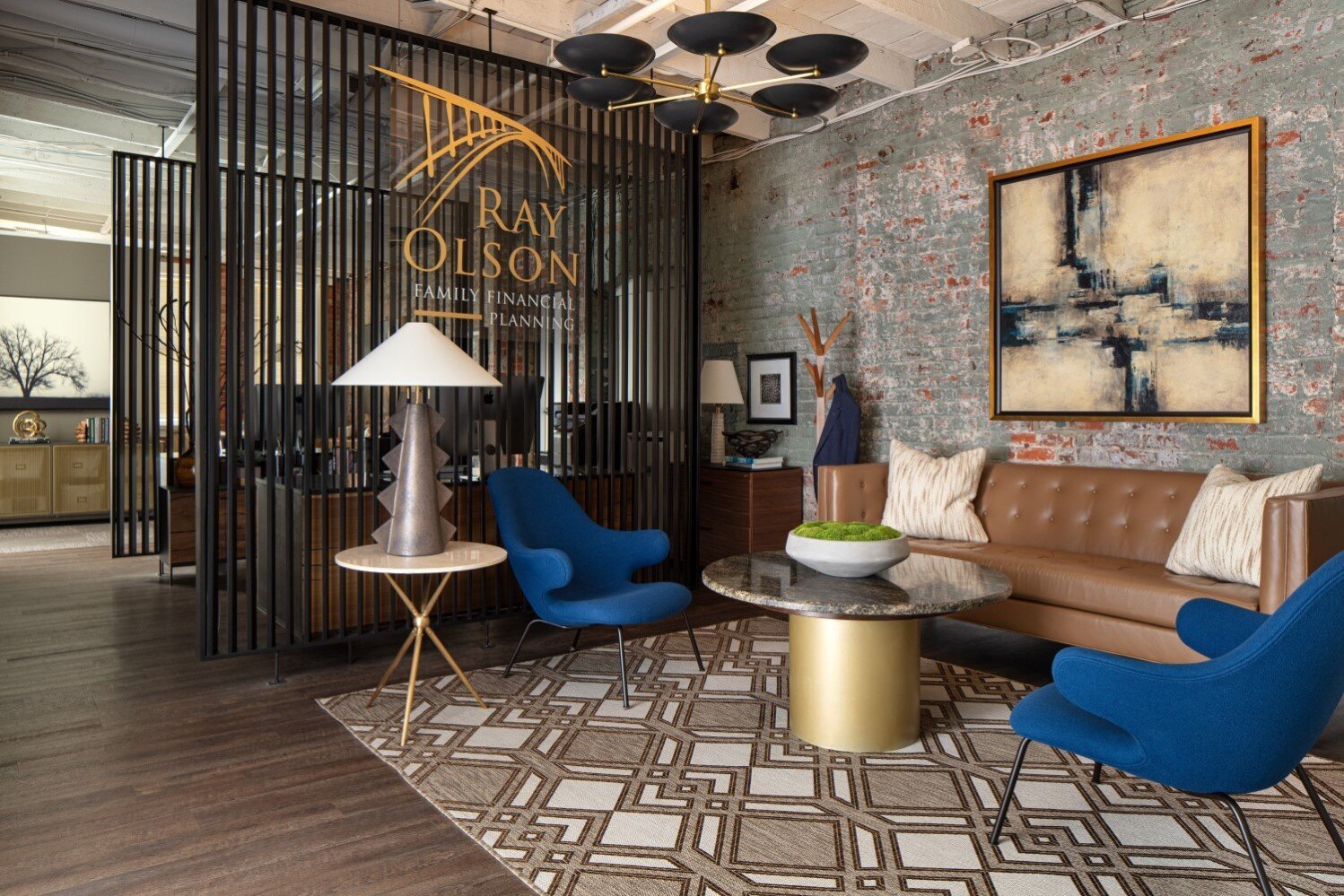 A modern office lobby with a leather sofa, two blue armchairs, a round marble table with a bowl of moss, and decorative lamp and artwork. The space features exposed brick walls and a glass partition with gold lettering for "Ray Olson Family Financial