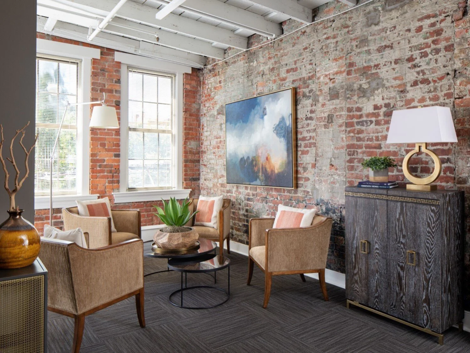 Living room with exposed brick wall, two large windows, a black coffee table with a green plant, four tan chairs with pink and white pillows, a dark wood cabinet with a white lamp, and a large abstract painting.