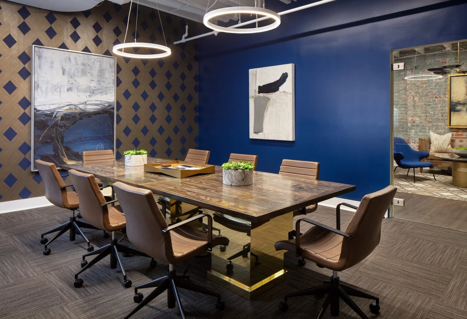 A modern conference room with a wooden table surrounded by eight tan leather office chairs on wheels. The room has a dark brown carpet, a wall with a blue accent color and art, and an adjacent room with exposed brick and contemporary furniture. Two c
