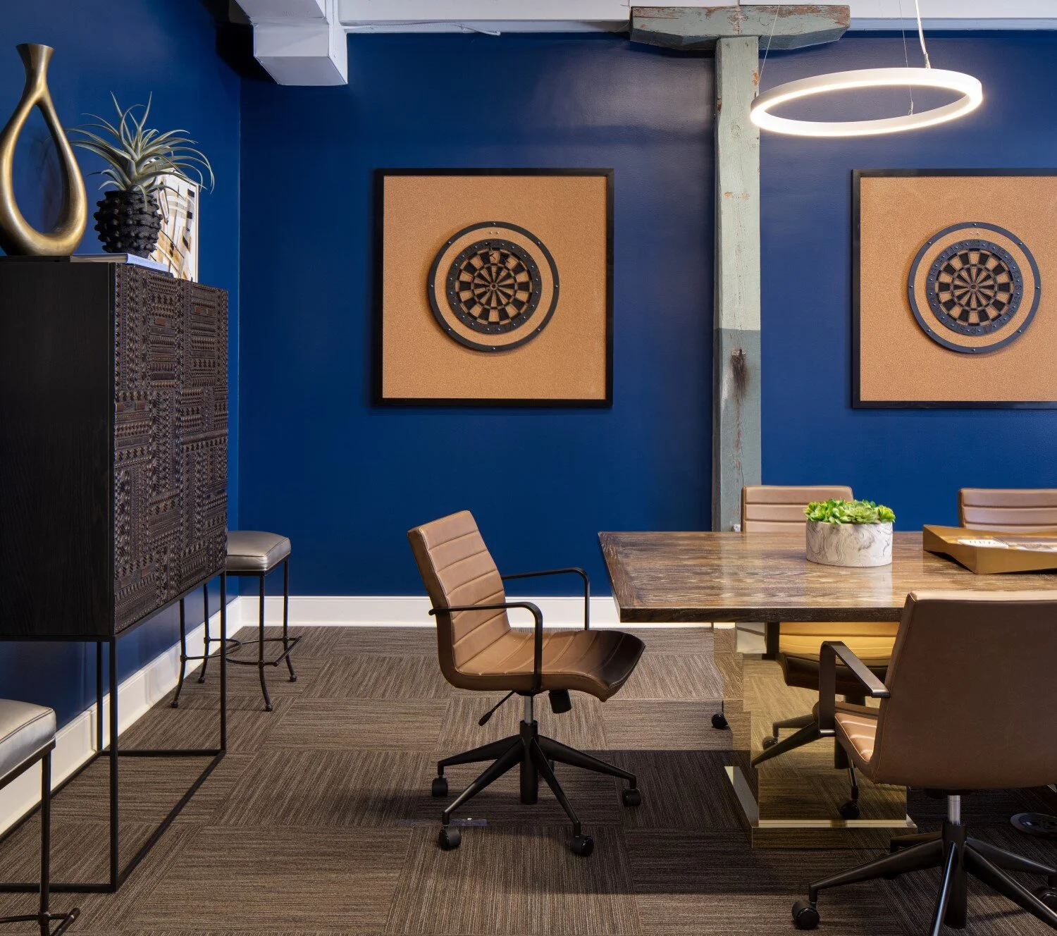 A modern conference room with a blue wall, framed dartboard art, a wooden table, and brown swivel chairs. Decorative vases and plants are on a black cabinet, and a circular ceiling light illuminates the room.