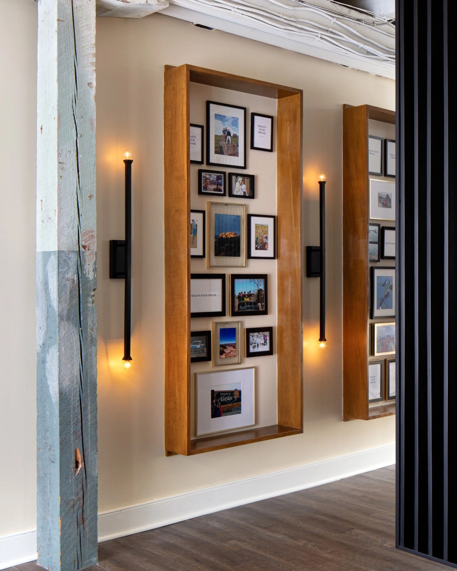 Wall display with a collection of framed photos and artwork, two modern black wall-mounted lights with Edison-style bulbs, wooden frames, and a beige wall in a contemporary interior.