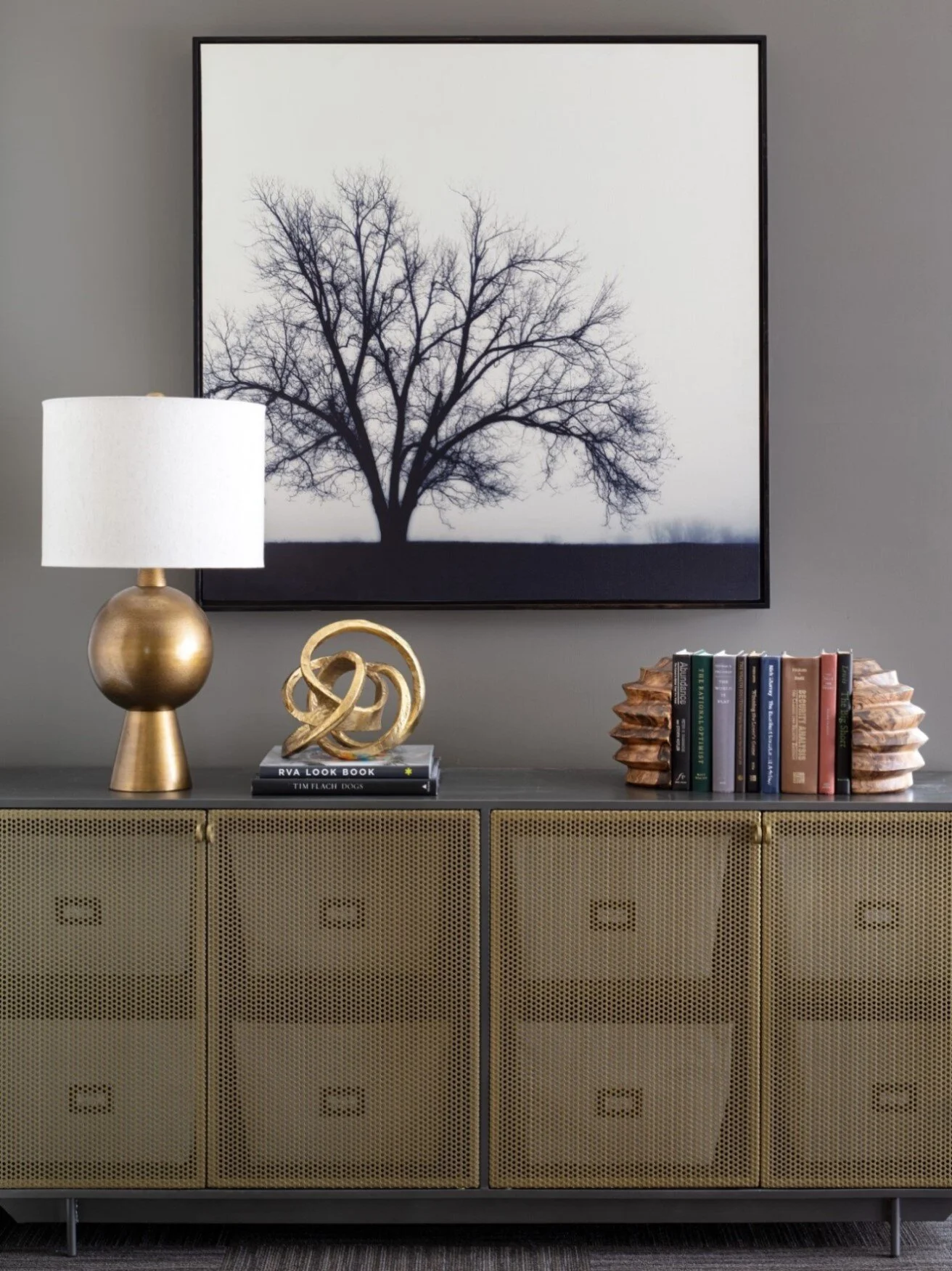Living room console with a framed black and white tree photograph, a gold table lamp with a white shade, a gold decorative sculpture, a stack of colorful books, and a metallic gold cabinet.