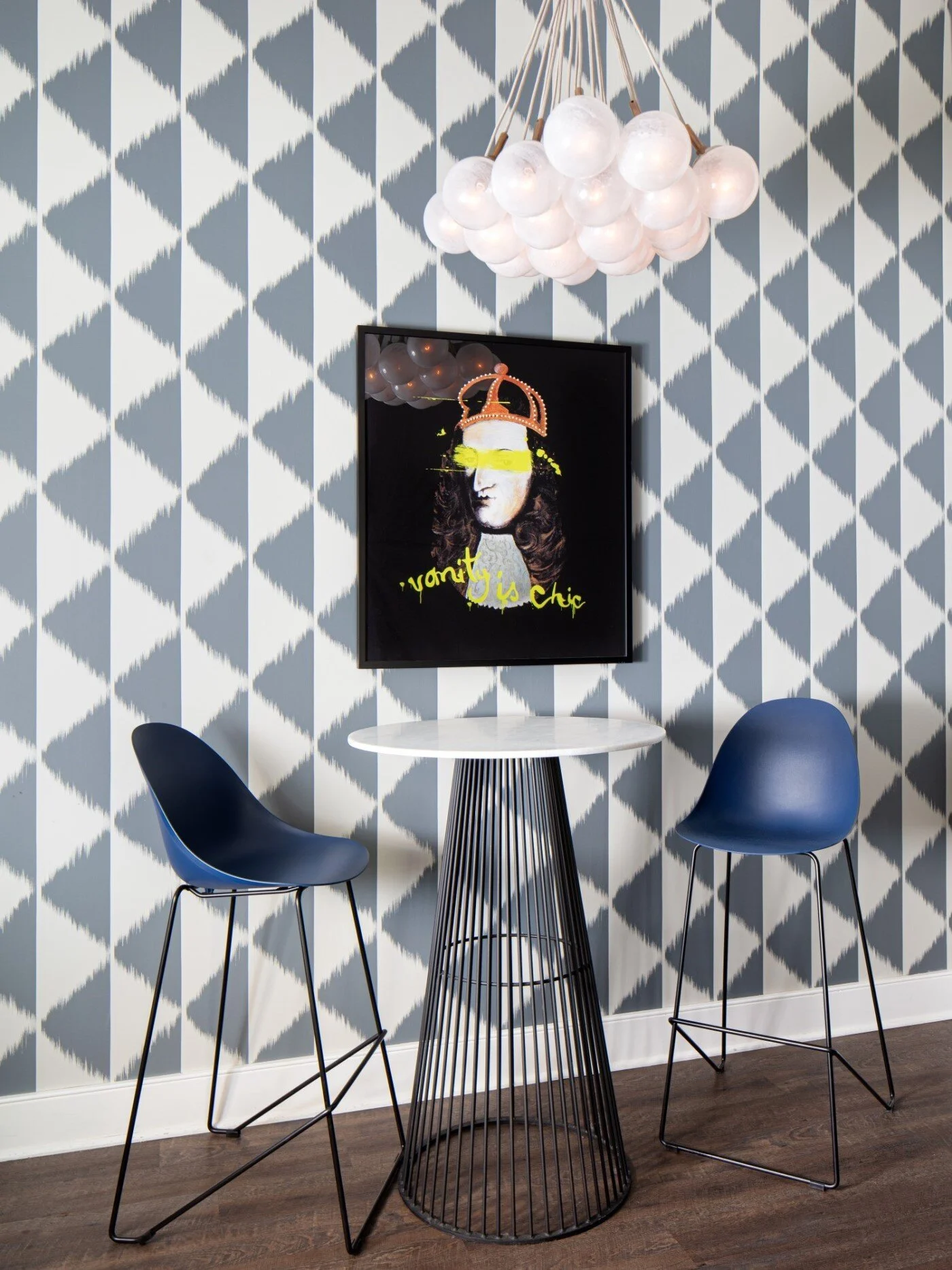 A modern interior space with a circular white table, two navy blue chairs with metal legs, a black wire-frame side table, a wall-mounted colorful art piece, and a white spherical chandelier hanging from the ceiling against a geometric patterned blue 