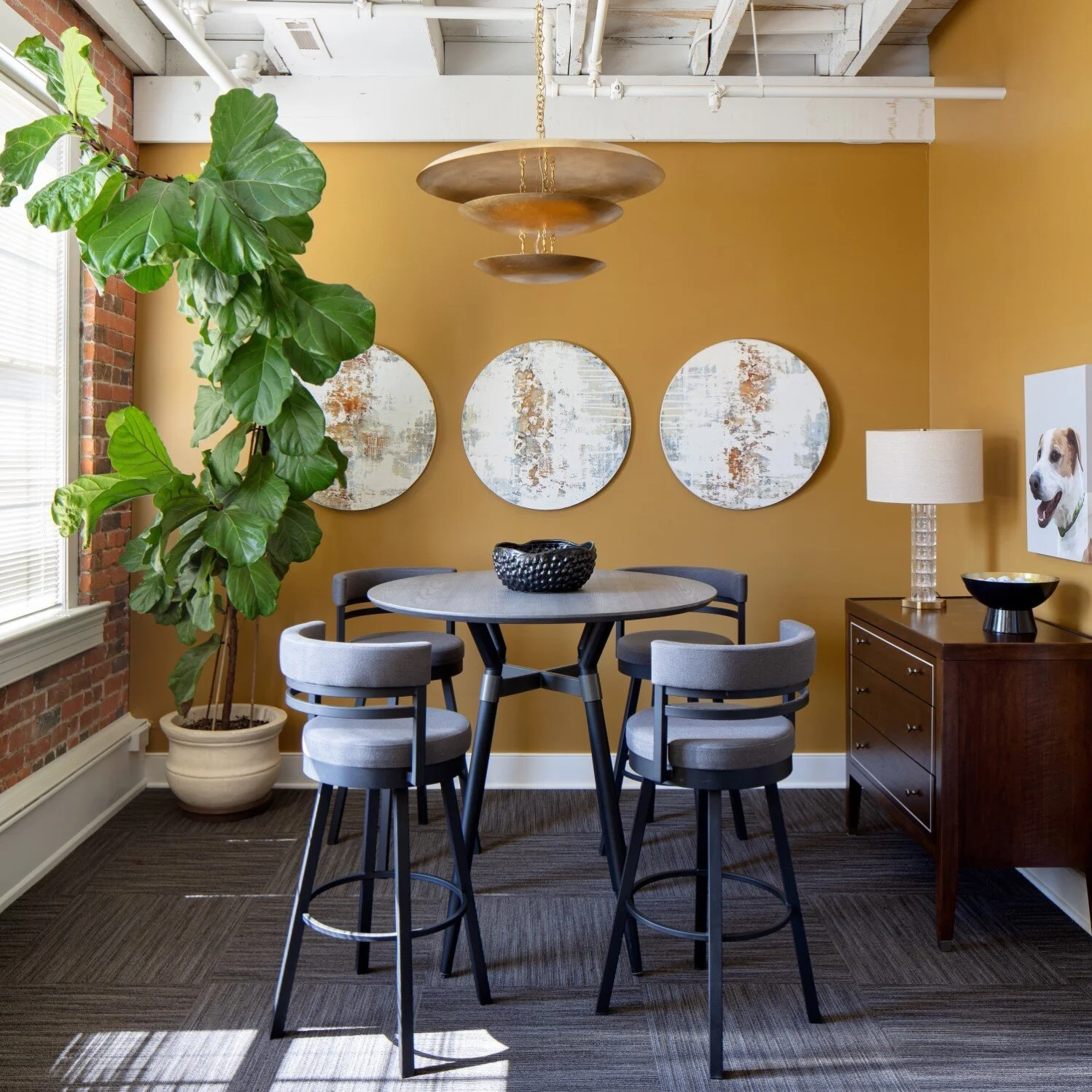 A dining area featuring a round table with four gray upholstered chairs, a large green potted plant, three round abstract paintings on a mustard-colored wall, a wooden sideboard with a lamp, and a window letting in natural light.