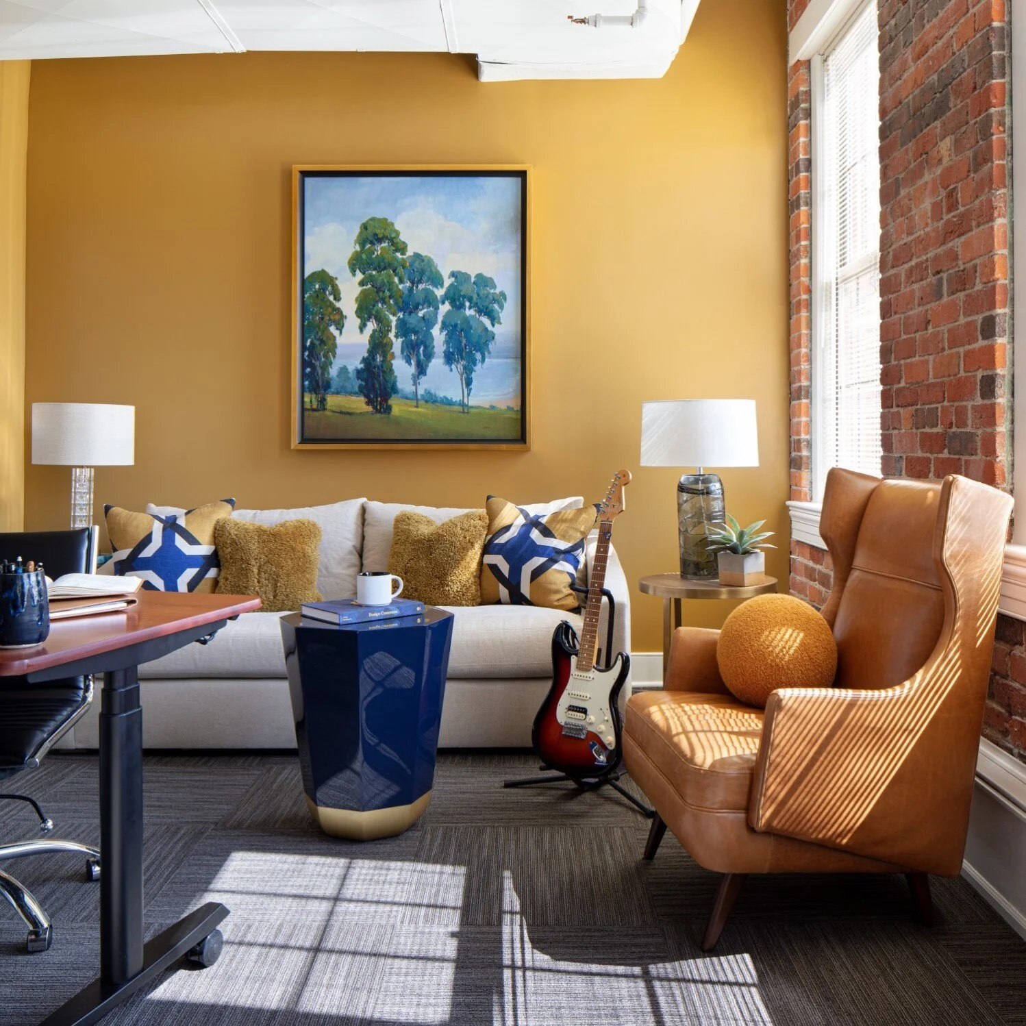 A cozy living room with a beige sofa, decorative pillows, a guitar, a side table with a plant and lamp, a leather armchair with a round pillow, a coffee table, and a colorful painting on a yellow wall. There are brick walls and windows letting in nat
