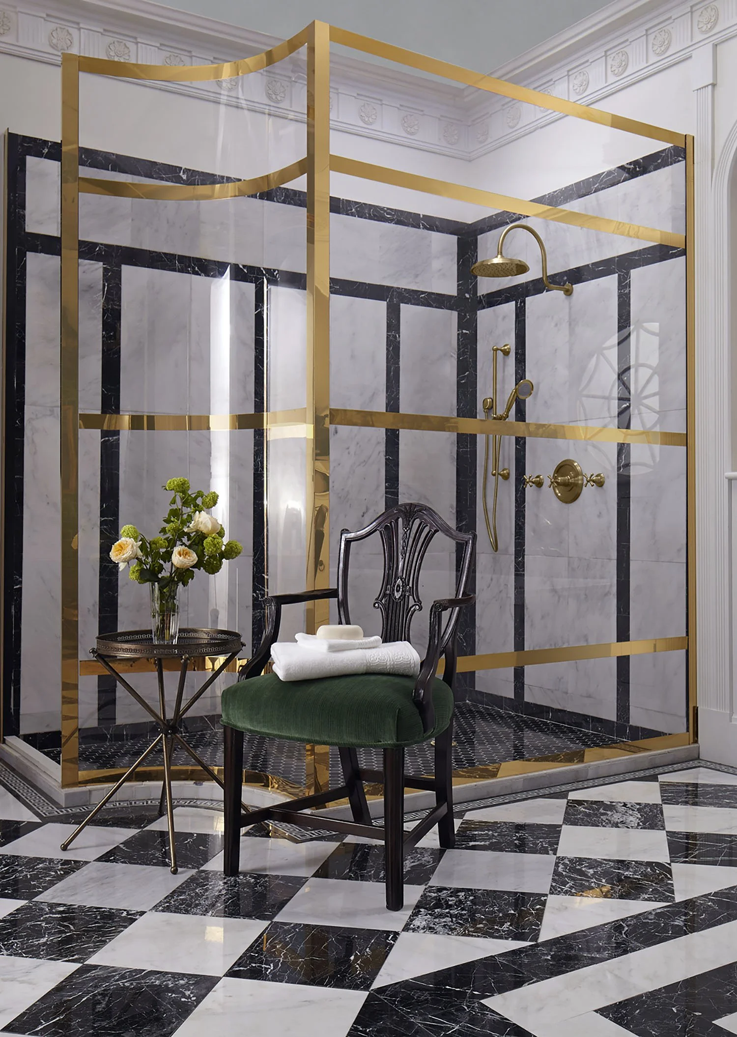 Luxurious bathroom with black and white marble tile flooring, ornate crown molding, and a glass shower enclosure with gold accents. A vintage-style black wooden chair with green upholstery sits next to a small round table with a vase of flowers and t