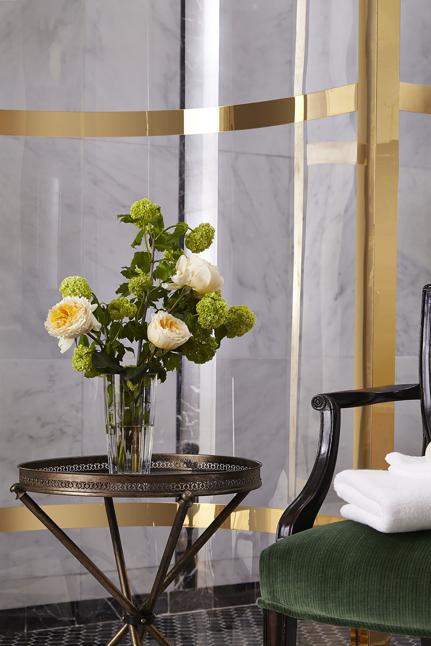 A bouquet of white and yellow flowers in a tall glass vase on a decorative metal table, next to a dark wooden armchair with green upholstery and white towels on the armrest, set against a marble wall with gold trim.