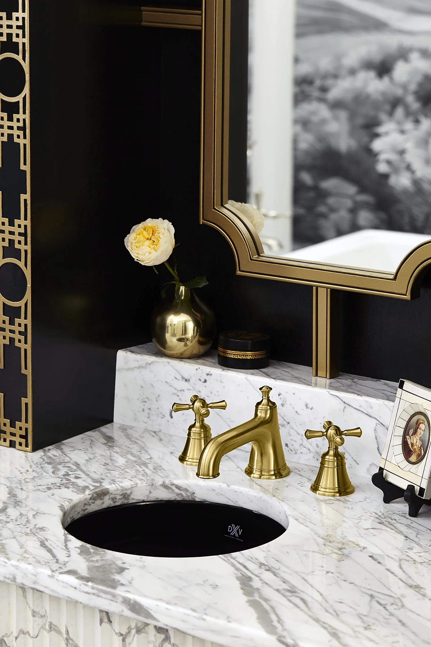 A luxurious bathroom vanity with a white marble countertop, a black round sink, and gold fixtures including a faucet and handles. There is a gold vase with a white flower, a framed picture of a woman, and a large mirror with a decorative gold frame.
