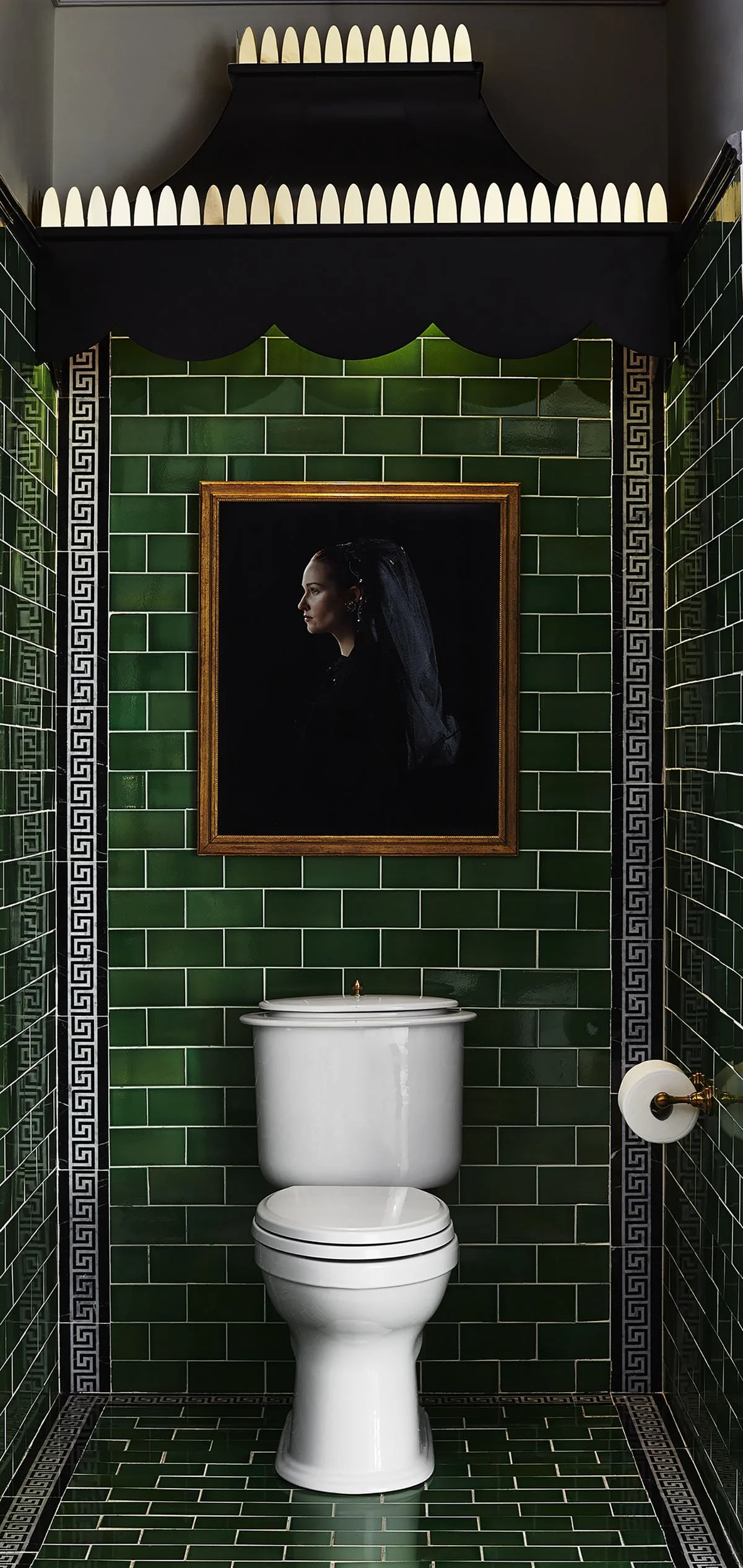 A room with green tiled walls and floor, a white toilet, a framed profile photograph of a woman with dark hair, and a decorative shelf above with white objects. There's also a mirror reflecting part of the room and a toilet paper holder on the right 