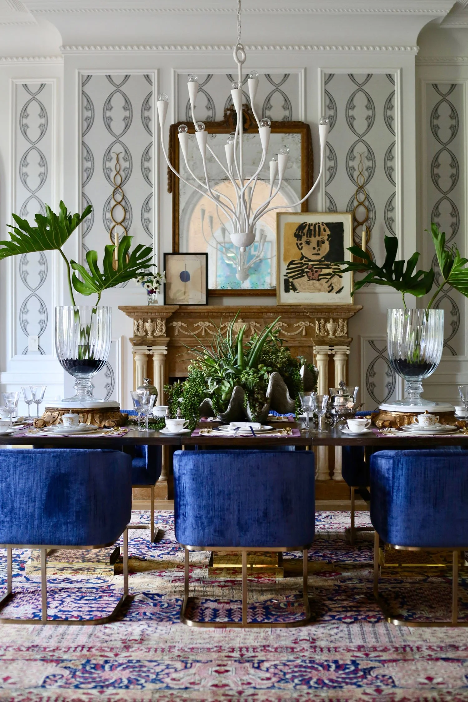Elegant dining room with a dark wood table set for a meal, surrounded by blue velvet chairs with gold legs. The table is decorated with large glass vases holding green monstera leaves, and a centerpiece of plants and rocks. A carved wooden mantel wit