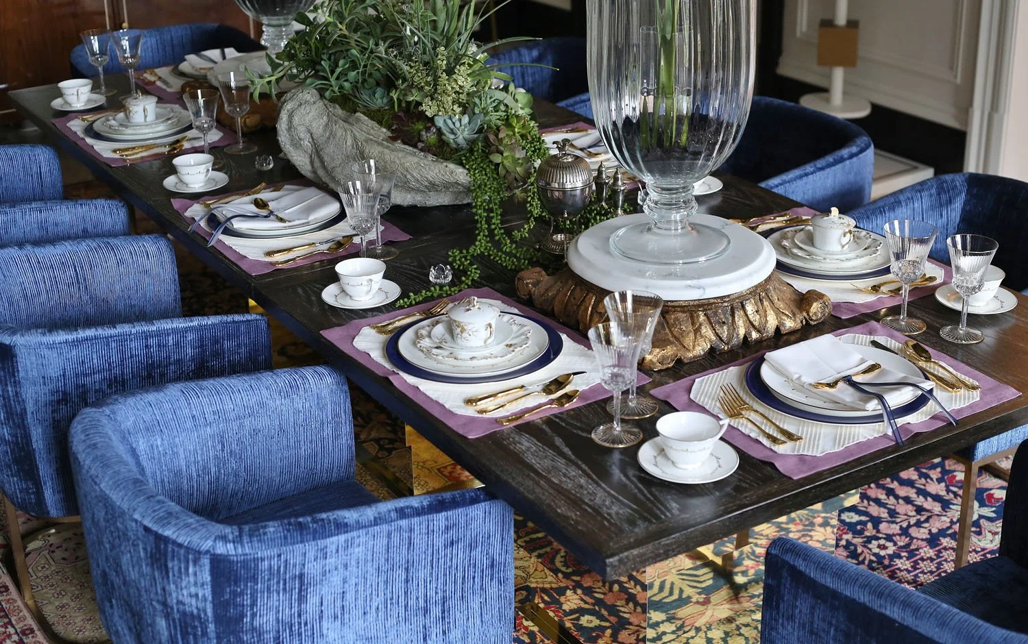 Elegantly set dining table with blue velvet chairs, white and gold china, purple placemats, and a large centerpiece with succulents and greenery, featuring a large vase in the middle.