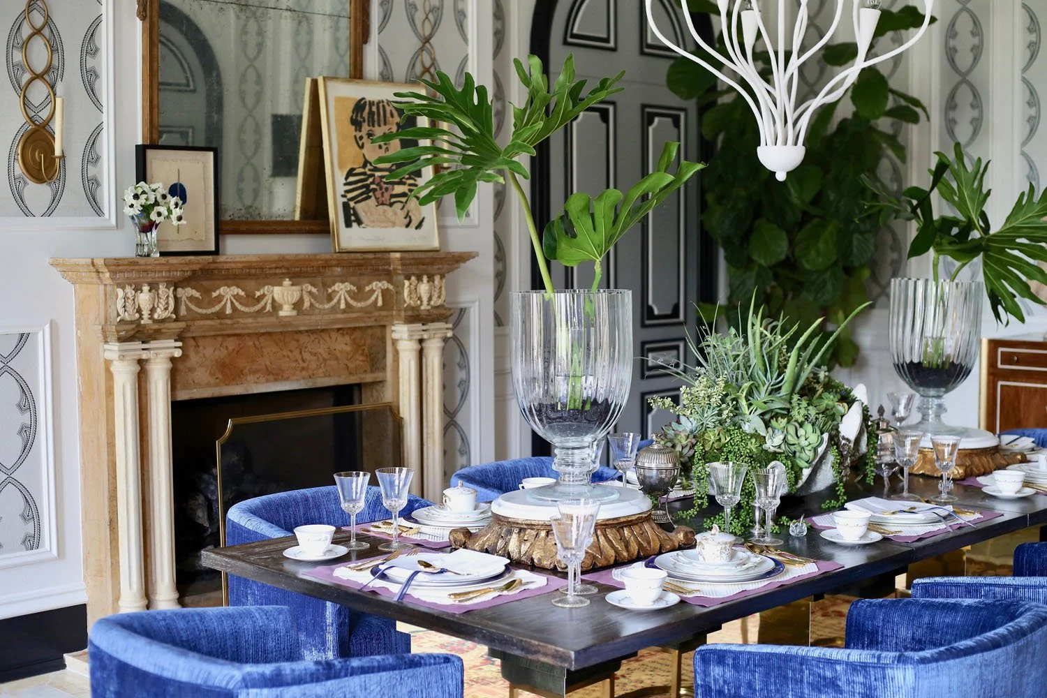 Dining room with a black table set with white china, gold cutlery, and purple napkins, surrounded by blue velvet chairs, a decorated fireplace with a mirror and artwork, and lush green plants as centerpieces.