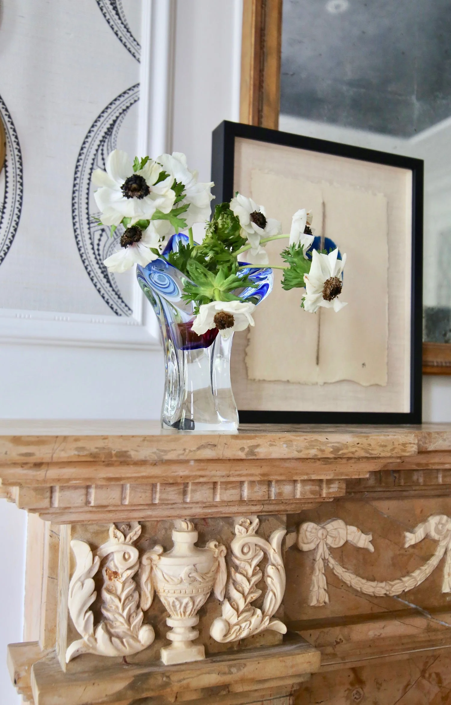 A clear glass vase with blue accents holding white flowers with dark centers, placed on an ornate wooden mantelpiece with carved details. Behind the vase, there is a framed piece of art with beige and black tones and a decorative wall segment with a 