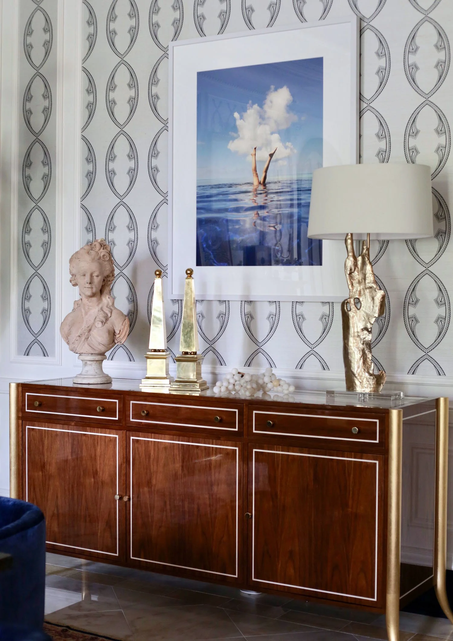 Elegant wooden sideboard with decorative items, including a classical bust sculpture, two geometric sculptures, a string of white beads, and a gold abstract lamp. A large framed photo of a person with legs in the water and clouds on the sky hangs on 