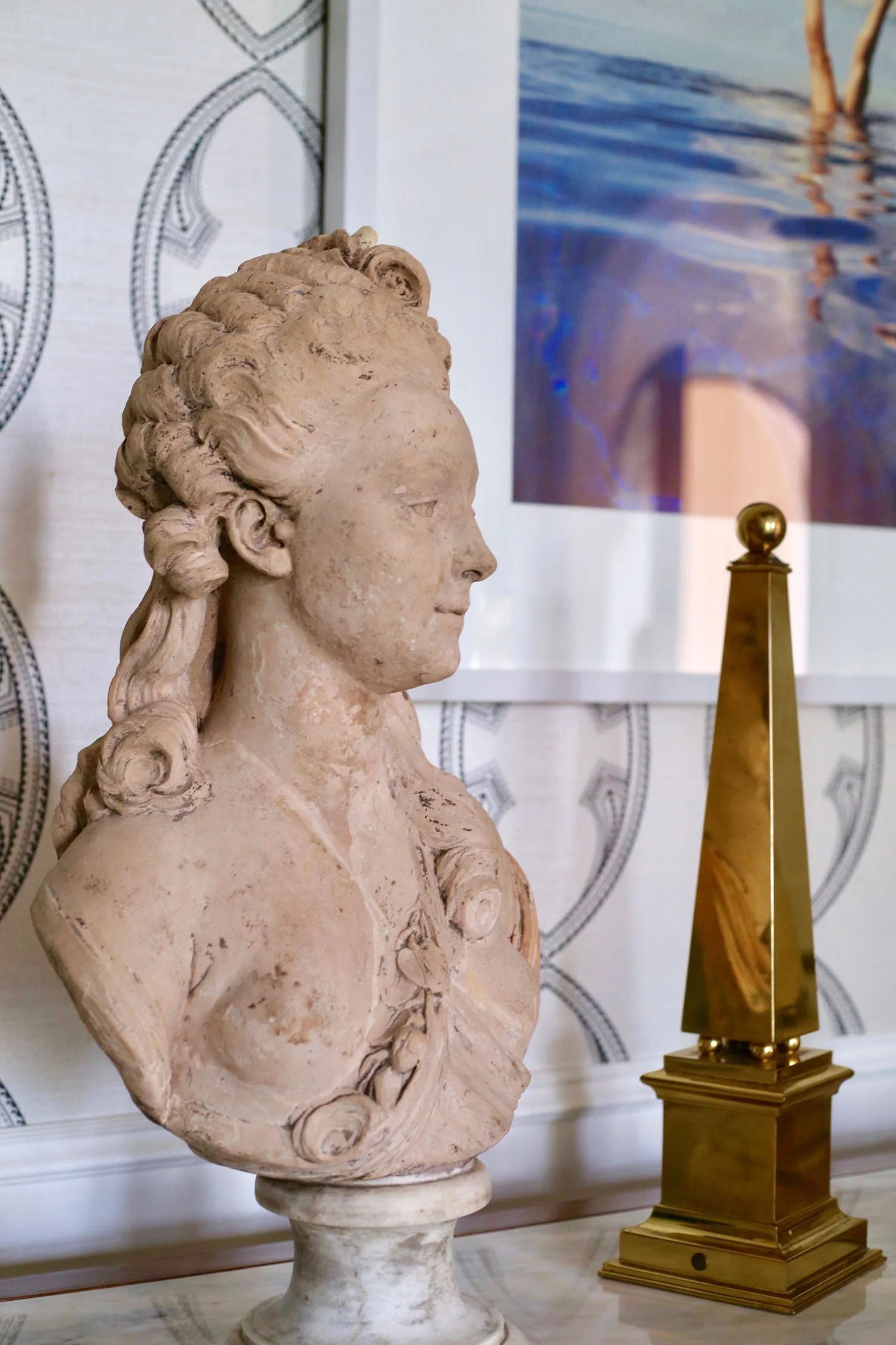 A classical marble bust sculpture of a woman with curly hair, placed on a pedestal, in a room with patterned wallpaper and artwork in the background.