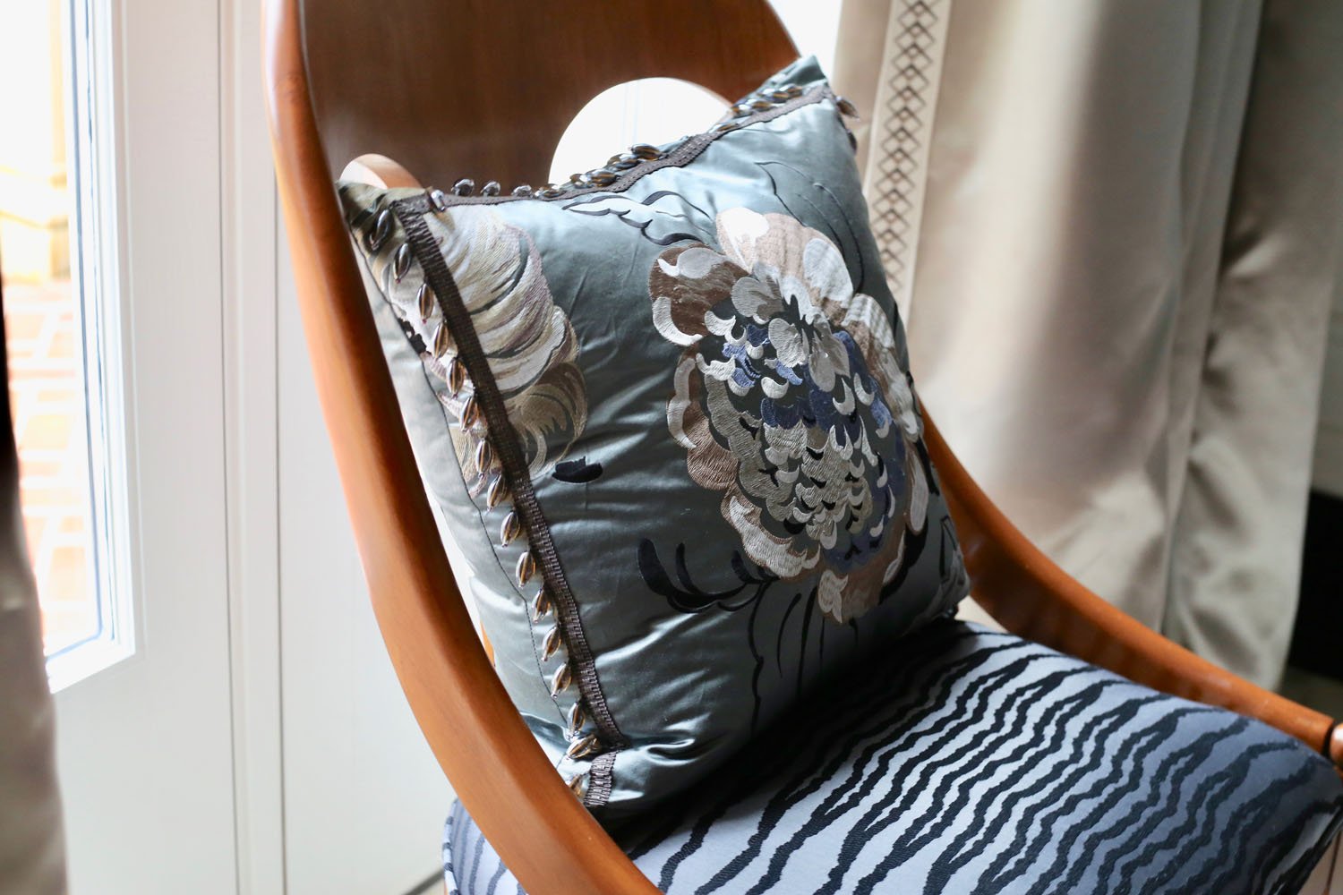 Close-up of a wooden chair with a decorative pillow featuring floral embroidery and a satin finish, placed near a window and curtain.