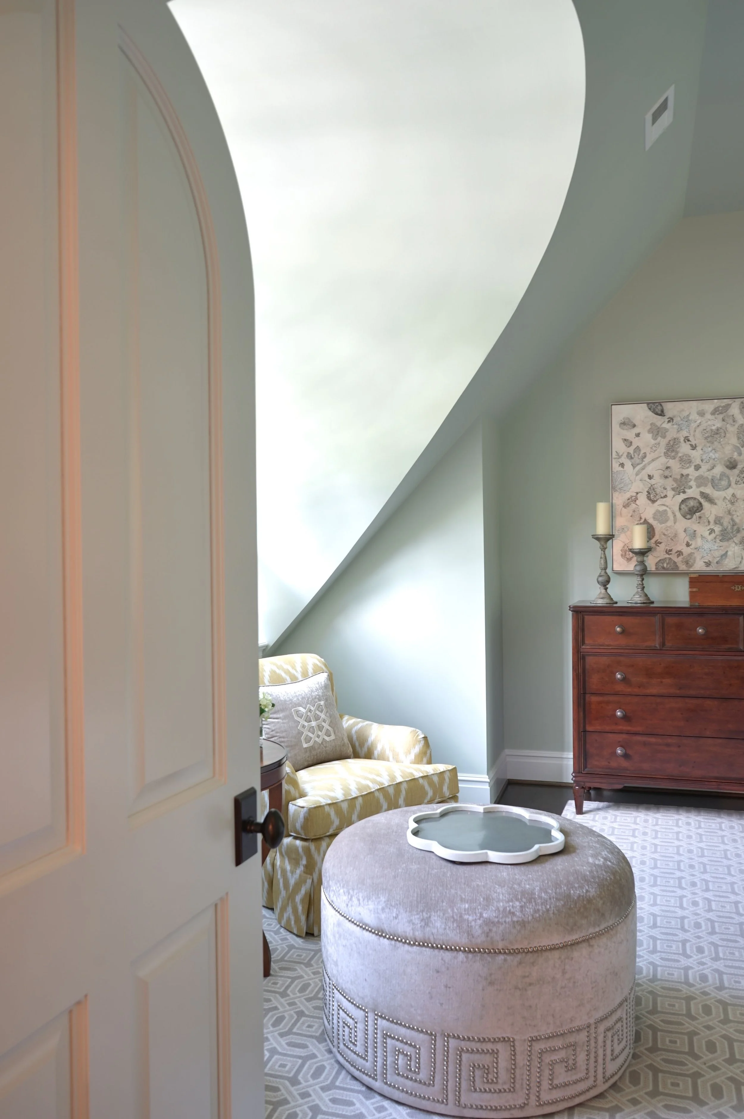 A room with accents of pastel green and beige, featuring a cushioned yellow and white floral sofa under a window with sloped ceiling, a wooden dresser with candles and a floral artwork, a round upholstered ottoman, and a patterned floor rug.