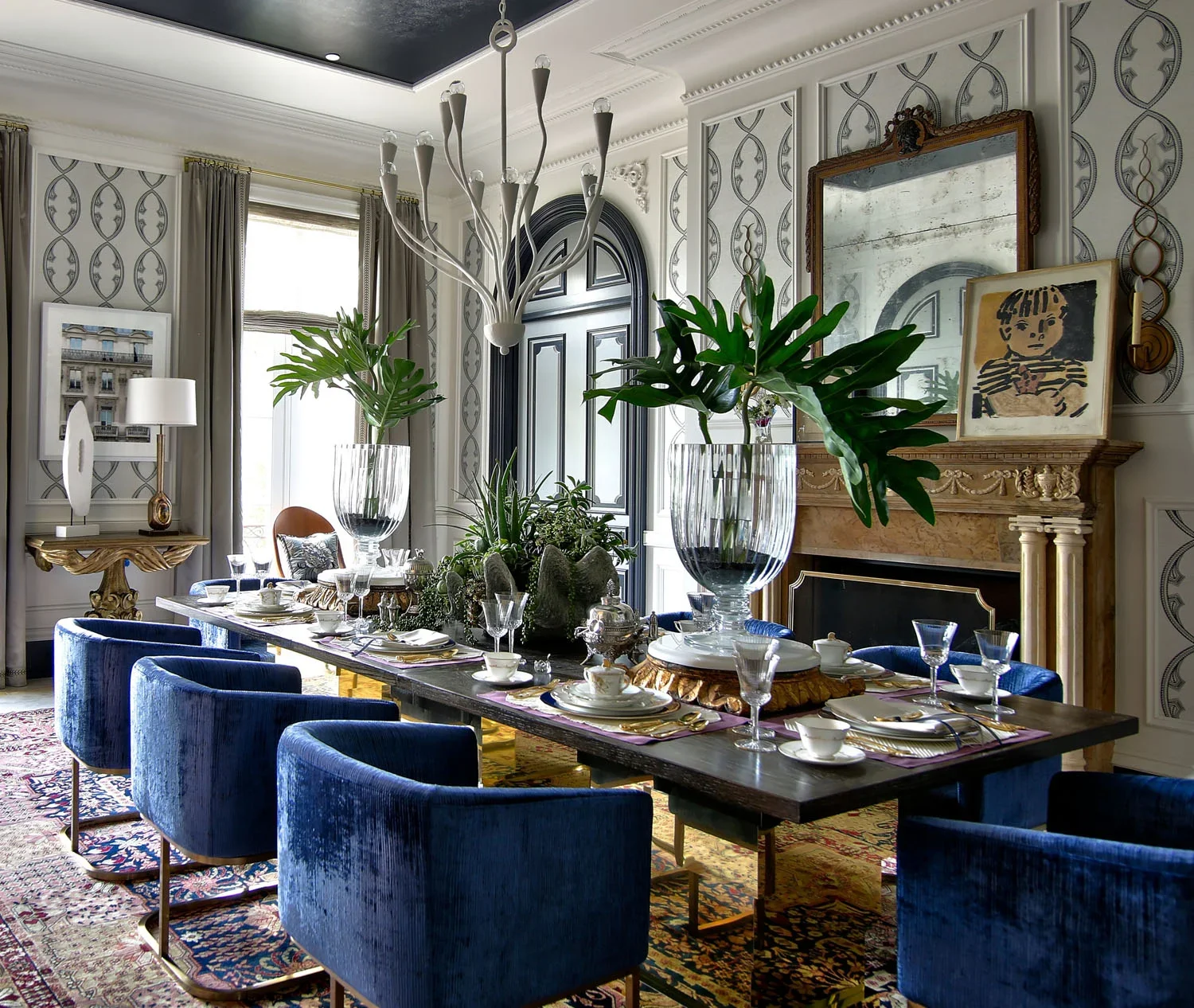 Elegant dining room with a long table set with fine china and glassware, surrounded by blue velvet chairs. The room features ornate wall panels, large windows with curtains, a chandelier, a fireplace with decorative items, and various framed artwork and mirrors.