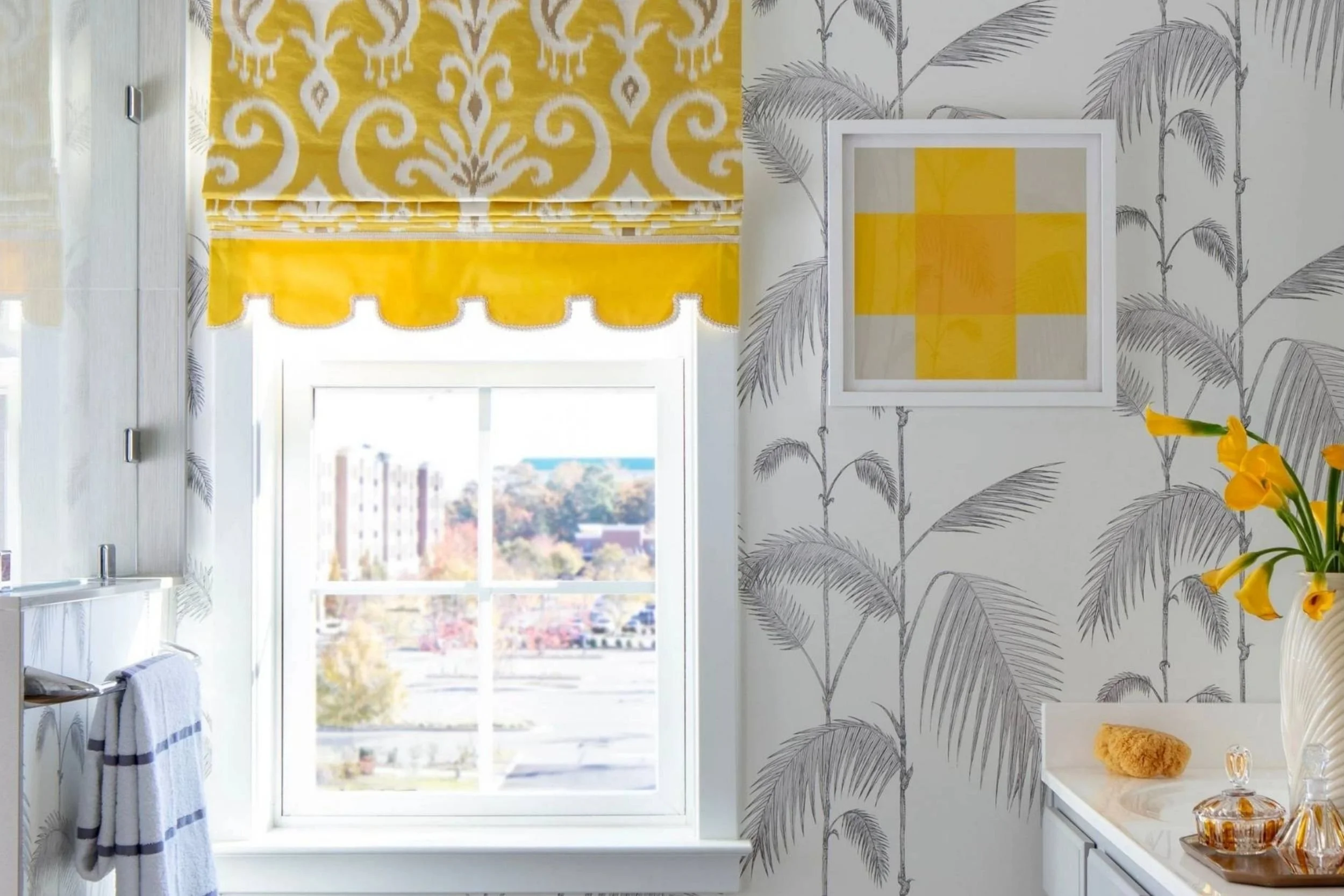 Yellow draperies and a small piece of yellow artwork contrast against a gray, wallpapered bathroom. Yellow flowers and yellow perfume bottles sit on the counter to the right.