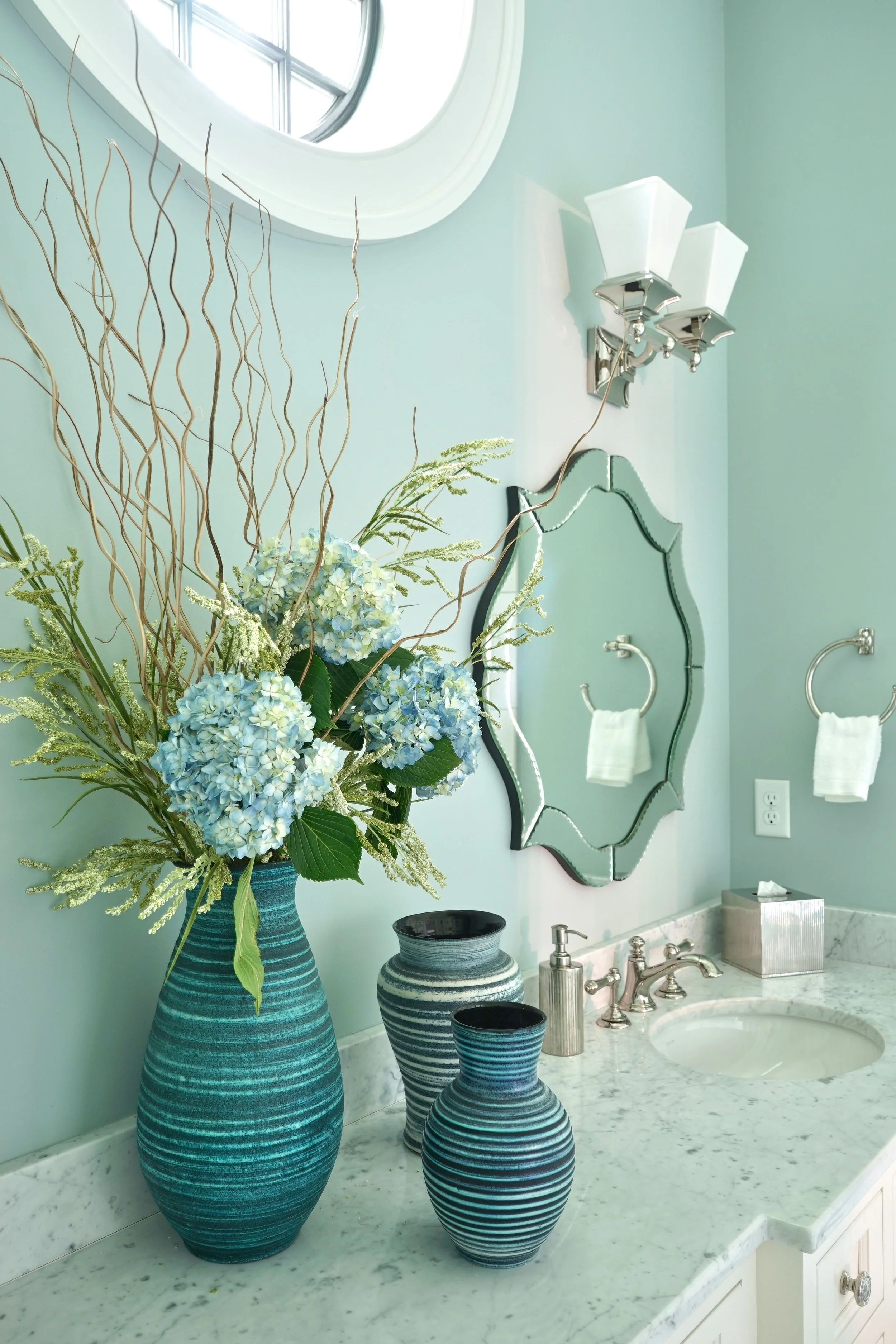 A bathroom countertop with three blue-striped vases containing a bouquet of white and blue hydrangeas and green foliage. There is a rectangular tissue box, a soap dispenser, and a pair of towels hanging on a mirror behind the countertop. The wall is 