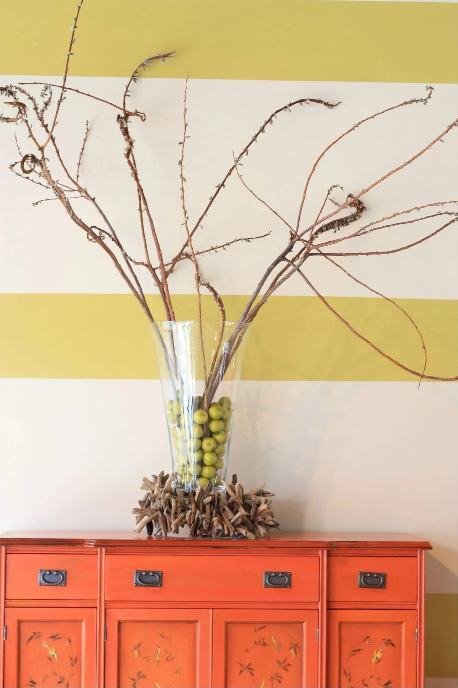 A clear vase with willow branches sits on an orange chest against a cream and yellow striped wall.