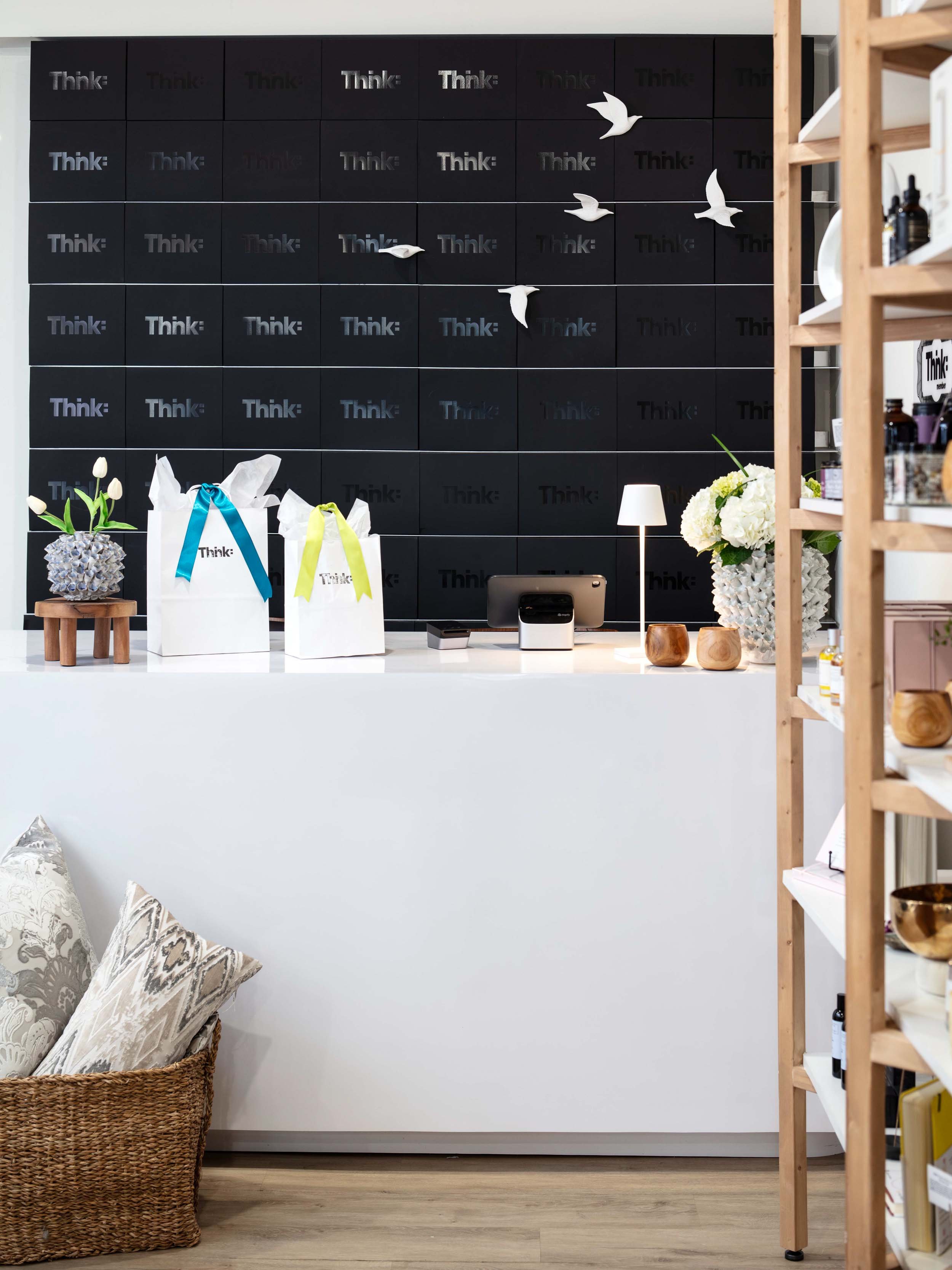 A modern reception desk with gift bags, vases of flowers, and a computer, set against a black wall with the word 'Think' and decorative white paper birds.