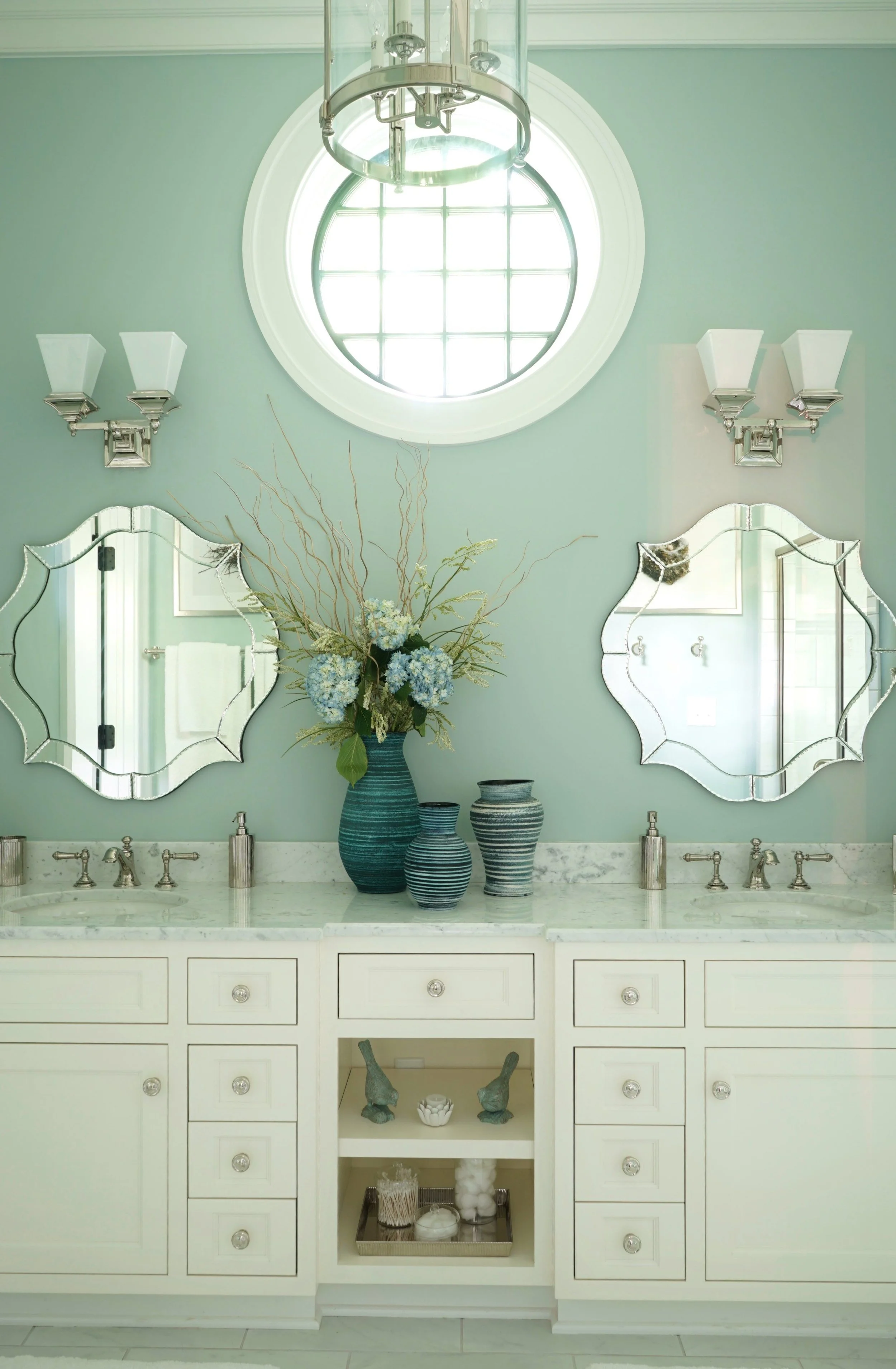 A bathroom vanity with a white cabinet, marble countertop, and silver fixtures. Two decorative mirrors are hanging above, along with two wall-mounted light fixtures. A large blue vase with floral arrangements sits on the counter, and various smaller 