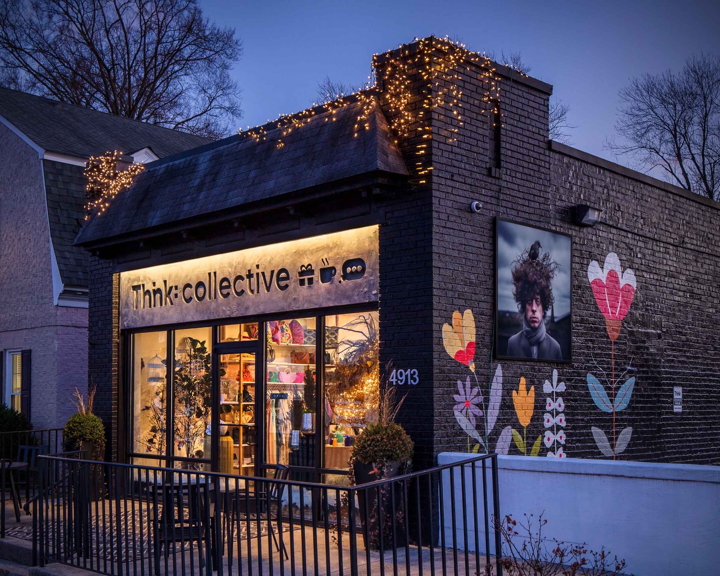 Storefront with illuminated sign reading 'Think: collective', decorated with string lights on the roof, featuring large window displays, painted flowers on the brick wall, and a large portrait of a young man with curly hair to the right of the entran