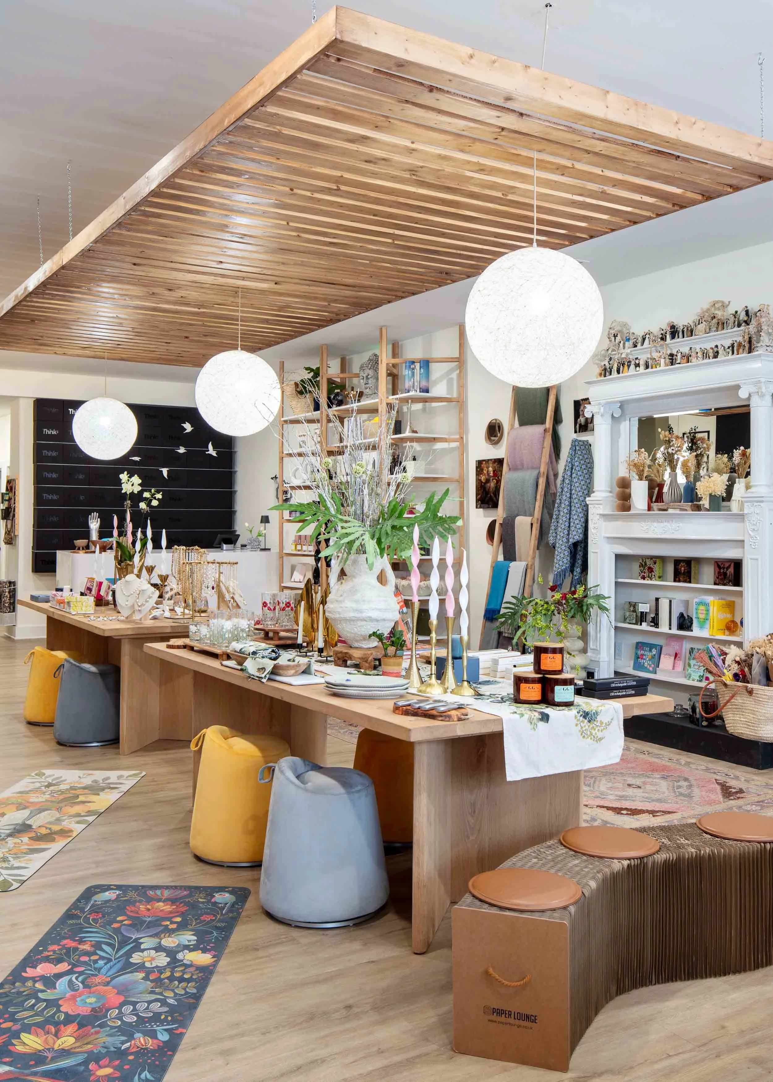 Interior of a boutique retail store with wooden ceiling and display tables filled with jewelry, candles, and decor items. Colorful rugs on the floor and a large floral arrangement in the center.